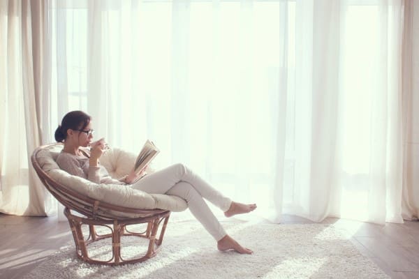 Frau sitzt vor hellem Wohnzimmerfenster und genießt ein Buch und einen Tee