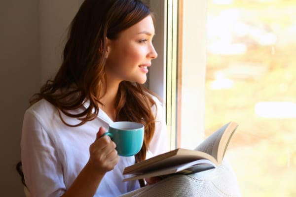 Frau mit Tee und Buch sitzt auf der Fensterbank eines wärmedämmenden Aluminiumfensters und schaut hinaus.