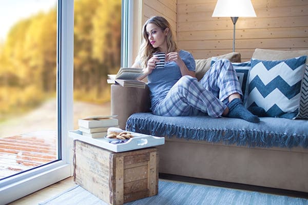Frau sitzt auf Couch und trinkt eine Tasse Tee und schaut dabei aus dem Fenster in eine Winterlandschaft. Frau sitzt auf Couch und trinkt eine Tasse Tee und schaut dabei aus dem Fenster in eine Winterlandschaft.
