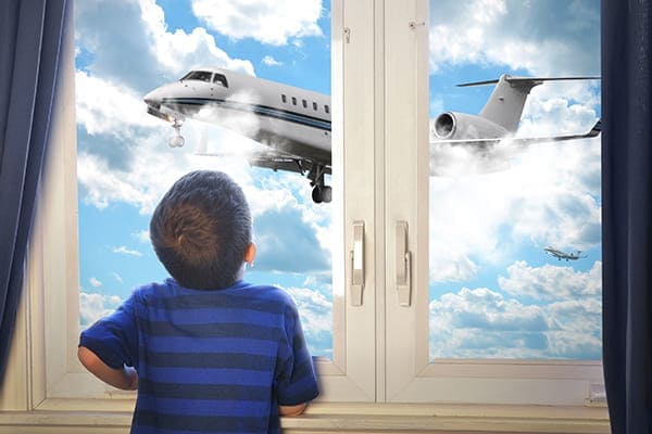 Kleiner Junge steht vor dem Fenster und schaut dem Flugzeug am Himmel zu.