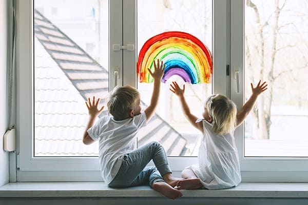 Kleine Kinder sitzen auf der Fensterbank und malen einen Regenbogen ans Fenster.