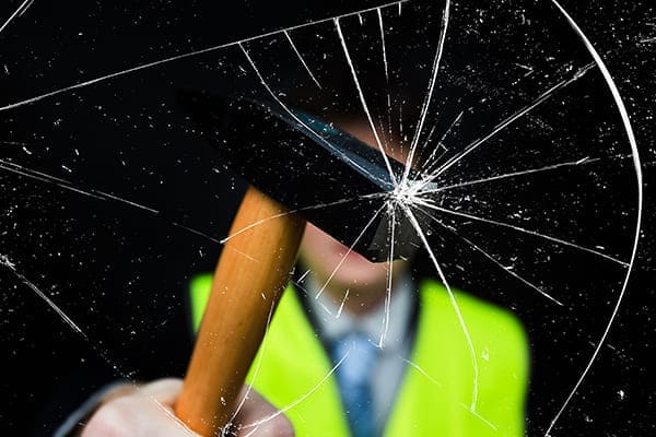 Mann mit gelber Weste im Hintergrund zerschlägt Glasscheibe mit einem Hammer. Mann mit gelber Weste im Hintergrund zerschlägt Glasscheibe mit einem Hammer.
