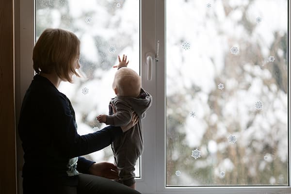 Frau hält kleines Kind das vor dem Fenster steht.