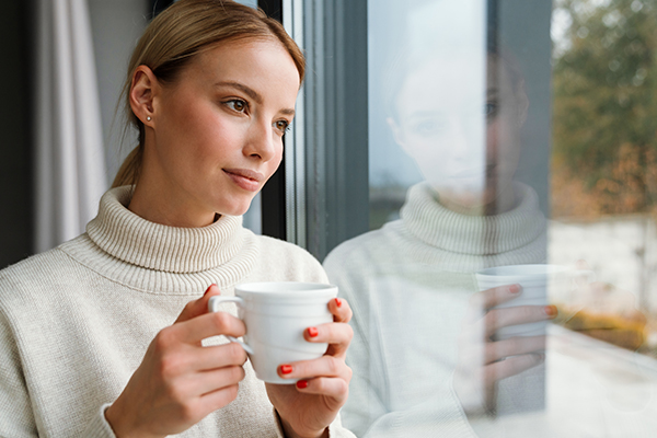Frau steht mit Tasse an der Scheibe und sieht nach draußen 